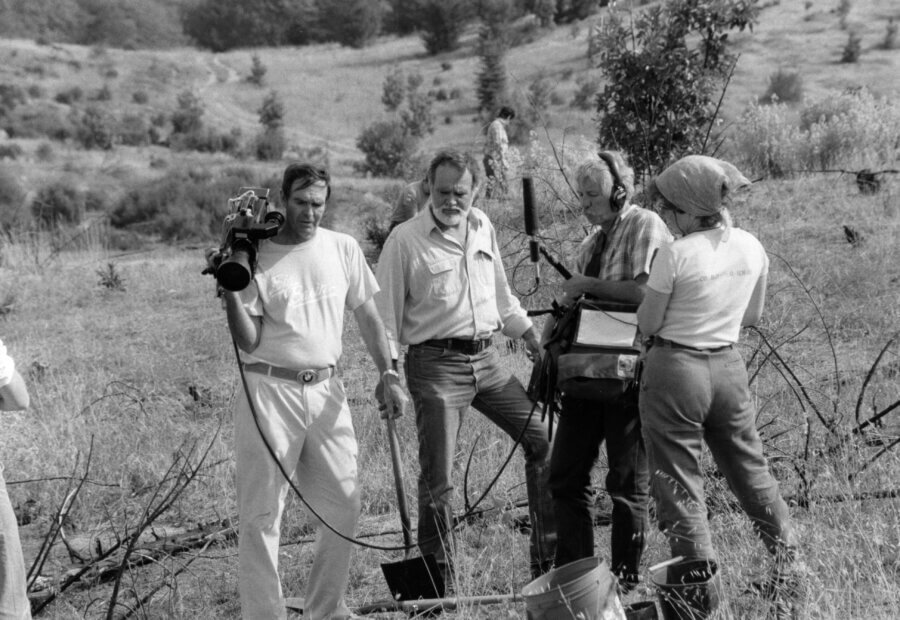 Four people stand in a grassy, rural field filming with a video camera and sound equipment. Two hold gear, one wears headphones, and one person faces away, all engaged in production work outdoors.