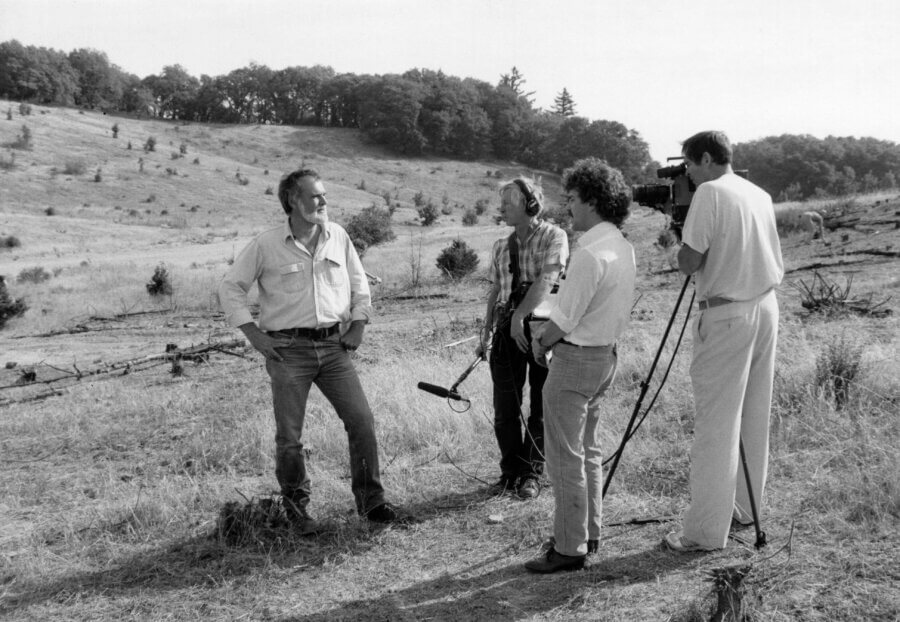 A person stands in a grassy field being interviewed by a small film crew with a camera, boom microphone, and sound equipment. Hills and trees are visible in the background.