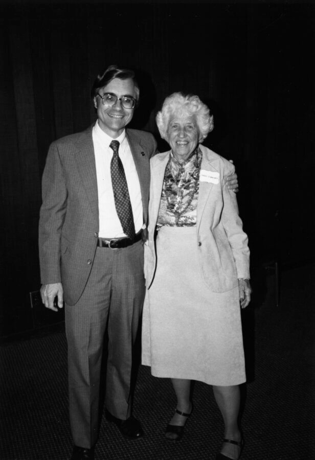 A person in a suit and tie stands smiling with an arm around another person in a skirt and jacket. They are both looking at the camera, standing indoors against a dark wall.