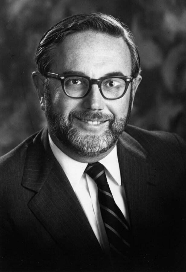 Black-and-white portrait of a person with glasses, a beard, and short hair, wearing a suit, white shirt, and striped tie, smiling and facing the camera against a dark background.