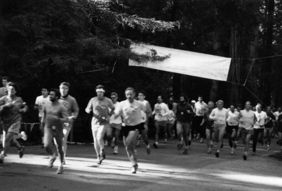 A group of runners, mostly in athletic wear, begin a race beneath a blank banner surrounded by tall trees in a forested area. The image is in black and white.
