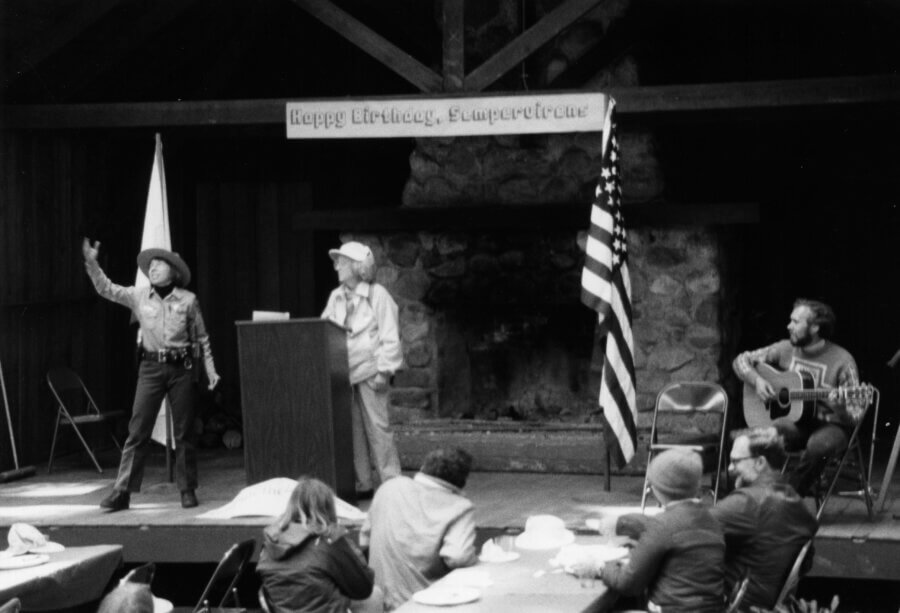 Black and white photo of people on a stage with a podium, U.S. flag, and a “Happy Birthday, Sempervirens” banner. One person gestures, another stands at the podium, and a third plays guitar while others watch from tables.
