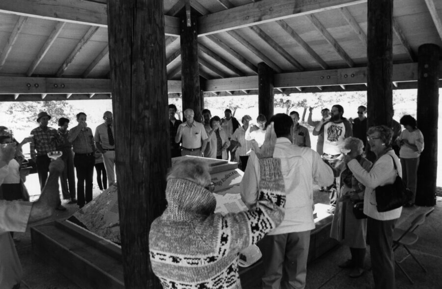 A group of people stand in a circle under a wooden pavilion, some with hands raised, around a covered object or model in the center. The scene appears to be a communal or ceremonial gathering.