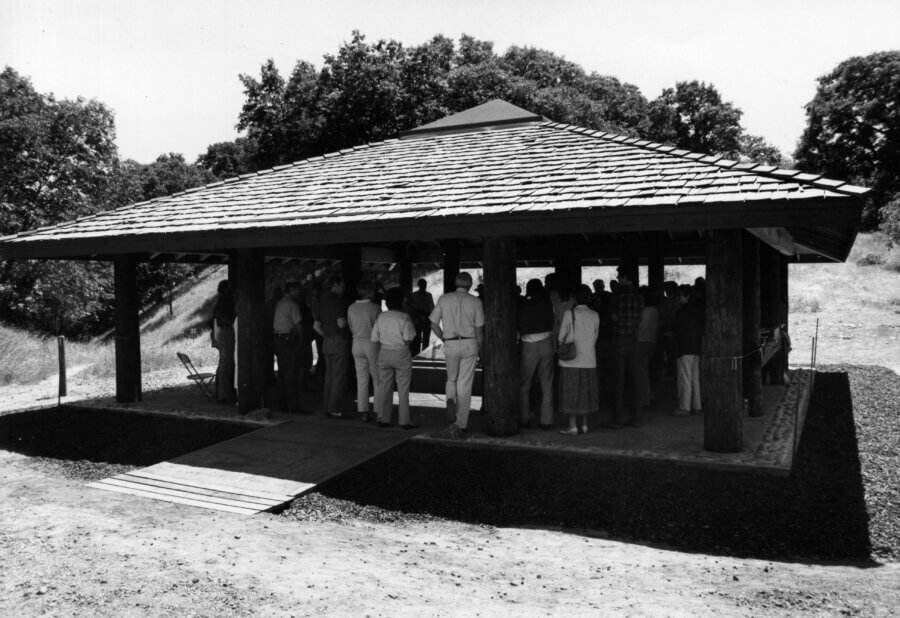 A group of people gather under a large, open-sided pavilion with a wooden shingle roof, surrounded by grassy hills and trees on a sunny day.