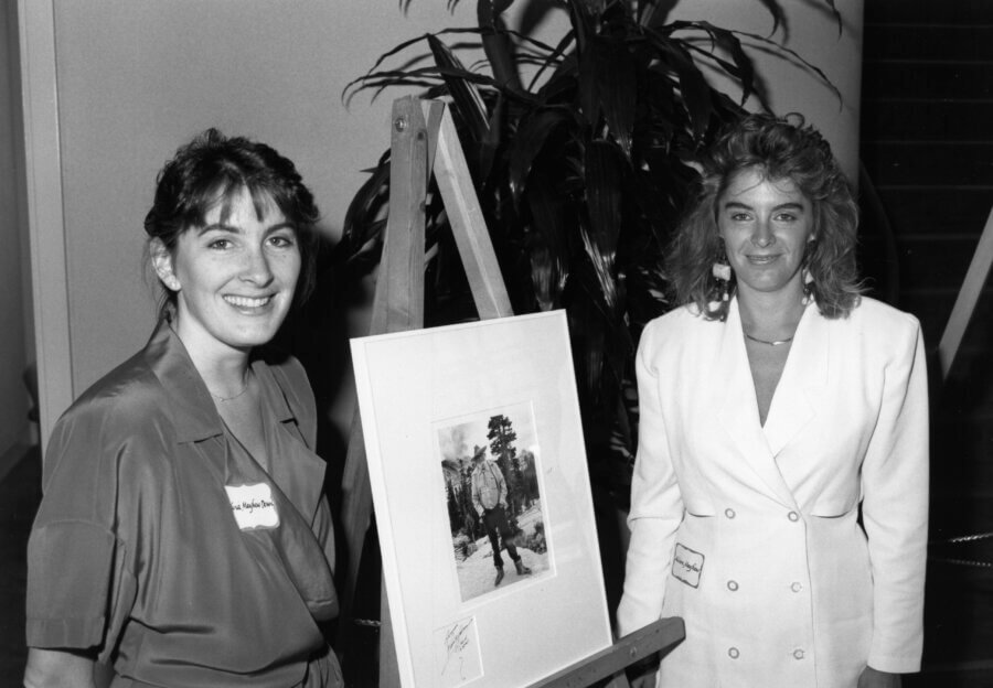 Two people stand beside an easel displaying a framed photo of a person outdoors. One person wears a dark outfit and smiles, while the other wears a white blazer. There is a large plant in the background.