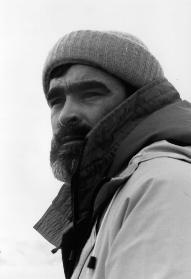 Black and white photo of a person wearing a knit beanie and a heavy jacket, looking thoughtfully into the distance. The background is plain and bright.