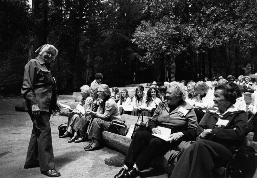 A group of people sit outdoors on benches in a wooded area, talking and holding papers, while one person stands and faces them; more people are seated in the background.