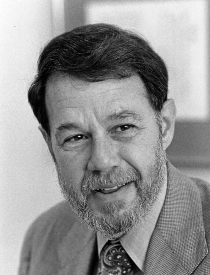 A grayscale portrait of a person with a beard and mustache, wearing a suit jacket, dress shirt, and patterned tie, smiling slightly and looking into the camera.