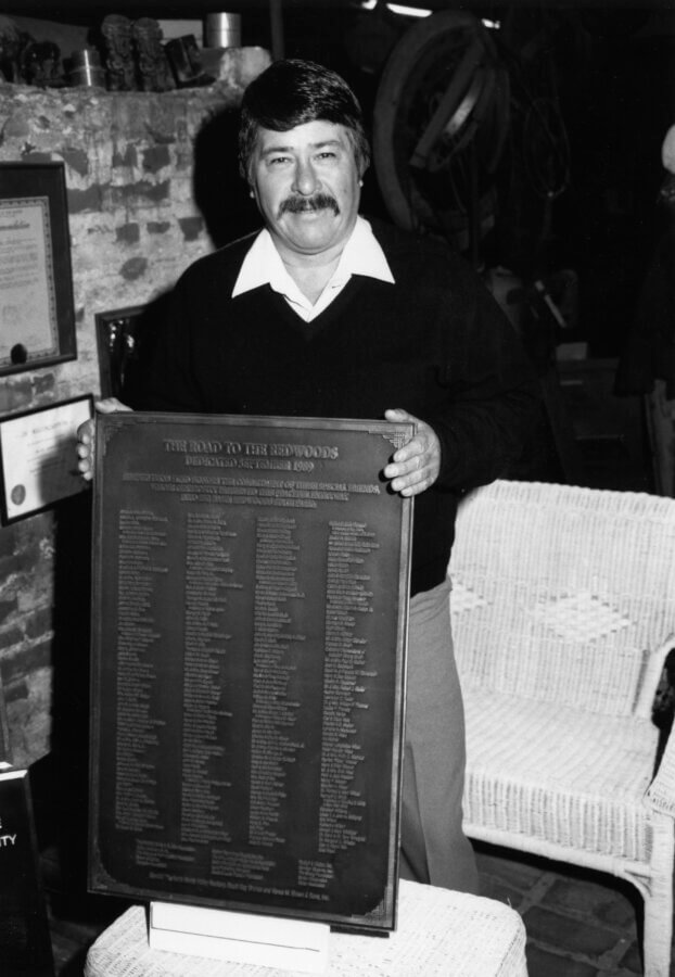 A person with a mustache stands indoors, holding a large plaque titled “THE ROAD TO THE REDWOODS” with many names engraved on it.