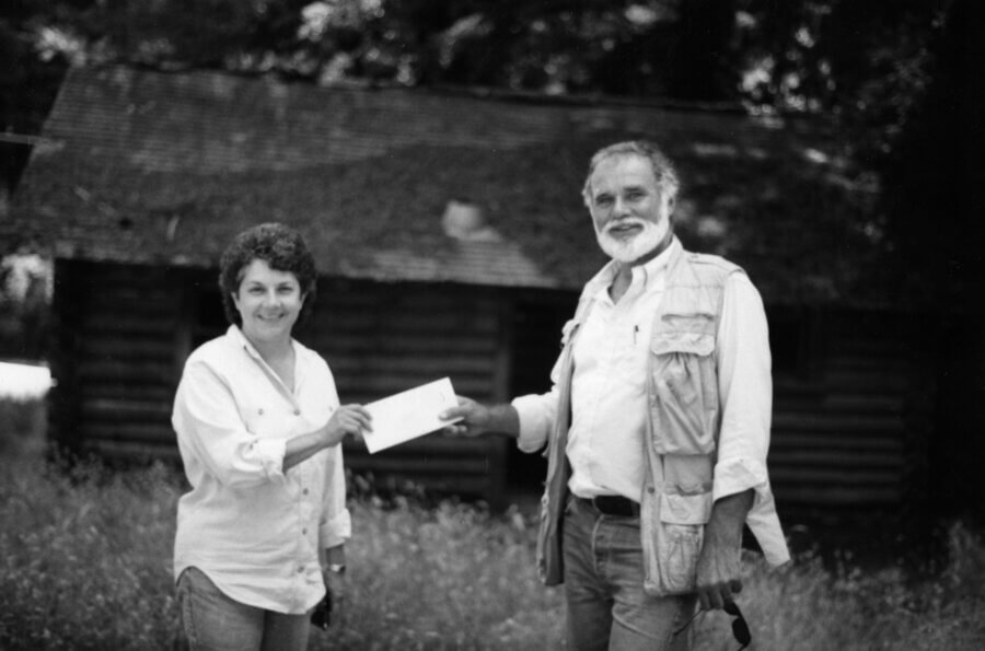 Two people stand outdoors in front of a rustic wooden cabin. One person is handing an envelope to the other person. Both are smiling and casually dressed. The scene appears to be in a grassy, wooded area.