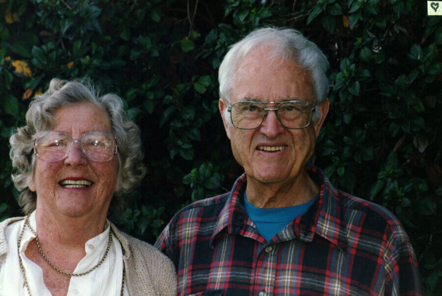 Two people smile while standing together outdoors, in front of leafy green shrubs. One wears glasses, a white blouse, and a beige sweater, while the other wears glasses and a red plaid shirt.