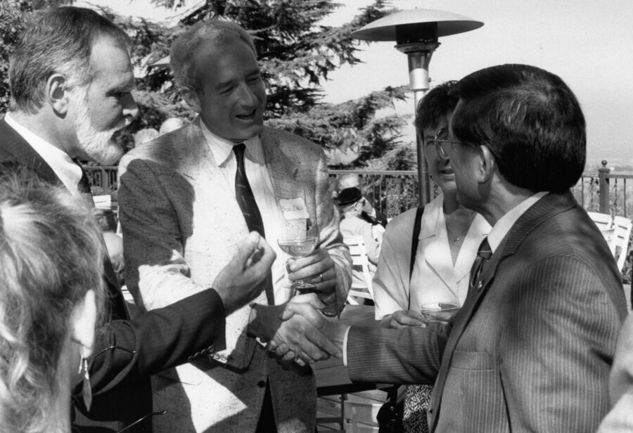 A group of people in formal attire are outdoors, engaging in conversation. Two people are shaking hands while others look on, smiling. Trees and a heater are visible in the background.