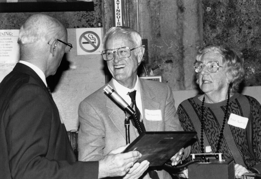 Three people stand together at an event. One person hands a plaque to another person, who is smiling.