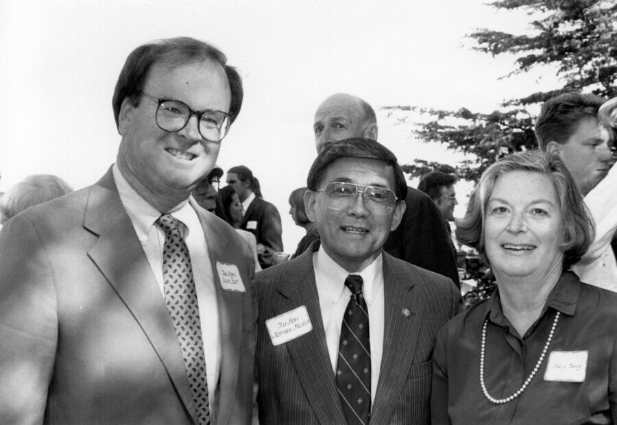 Three people smile at an outdoor event with other people in the background. All three wear name tags. Trees and a bright sky can be seen behind them.