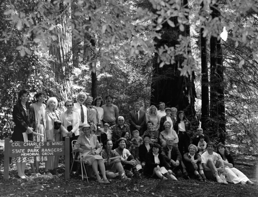 A large group of people, of various ages, poses together in a forested area near tall trees. In front, a sign reads “Col. Charles B. Wing State Park Rangers Memorial Grove.”.