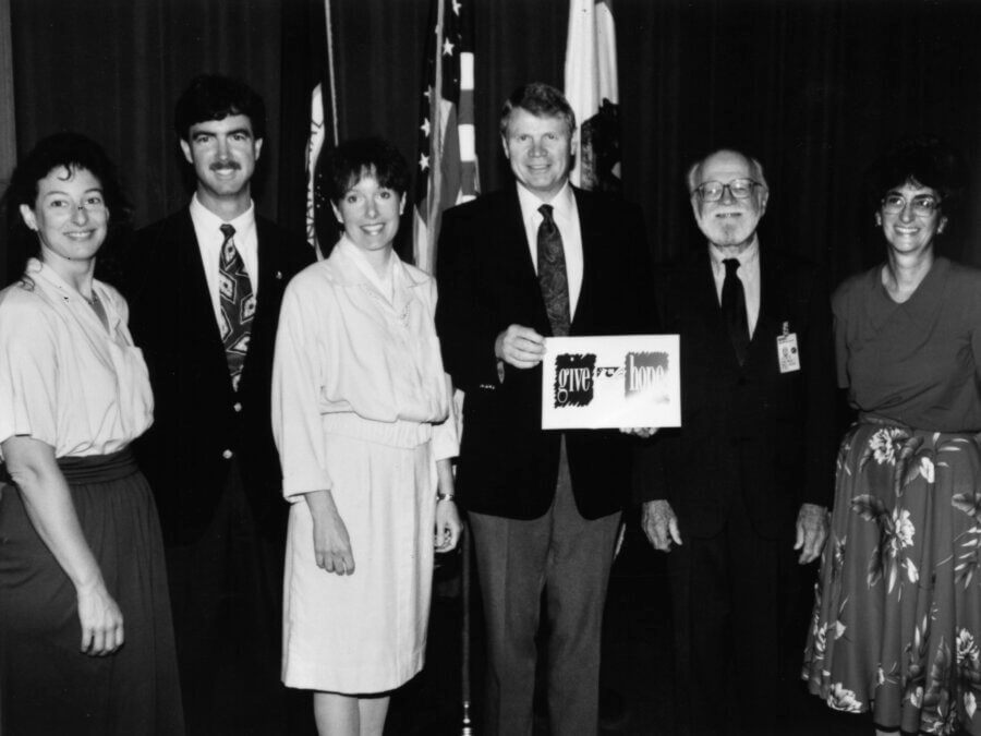 Six people stand in a row, dressed in business or semi-formal attire, in front of flags. The person in the center holds a plaque. All are smiling, and the photo is black and white.