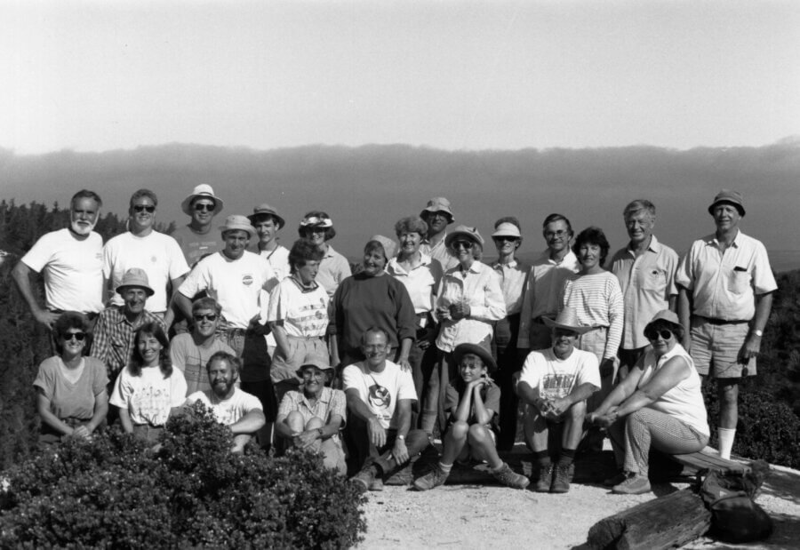 A group of about 25 people pose outdoors on a dirt path surrounded by greenery, with trees and a cloudy sky in the background. Most are smiling and dressed casually. The photo is black and white.
