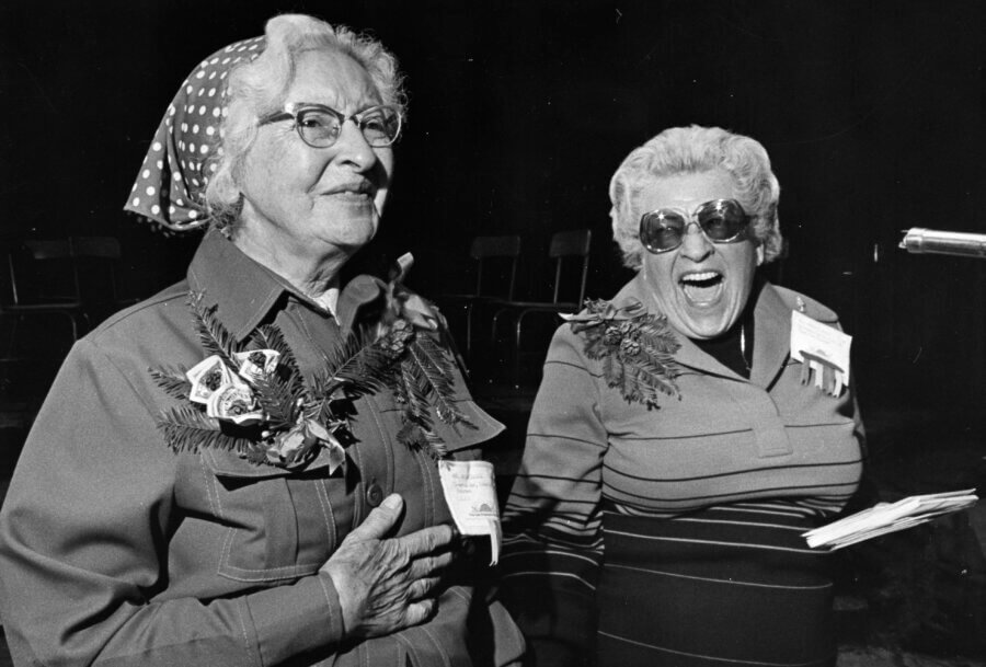Two people stand side by side, both smiling. One wears glasses, a polka-dot headscarf, and a flower corsage; the other wears sunglasses, a striped top, and holds papers, laughing exuberantly.