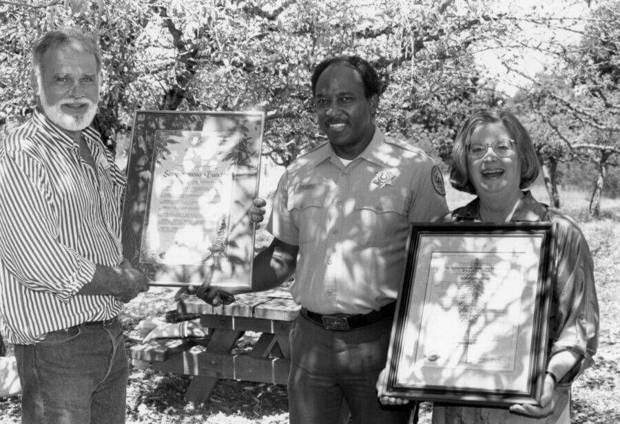 Three people stand outdoors under leafy trees, smiling and holding framed certificates or awards. Sunlight and shadows filter through the branches, and a wooden picnic table is visible in the background.