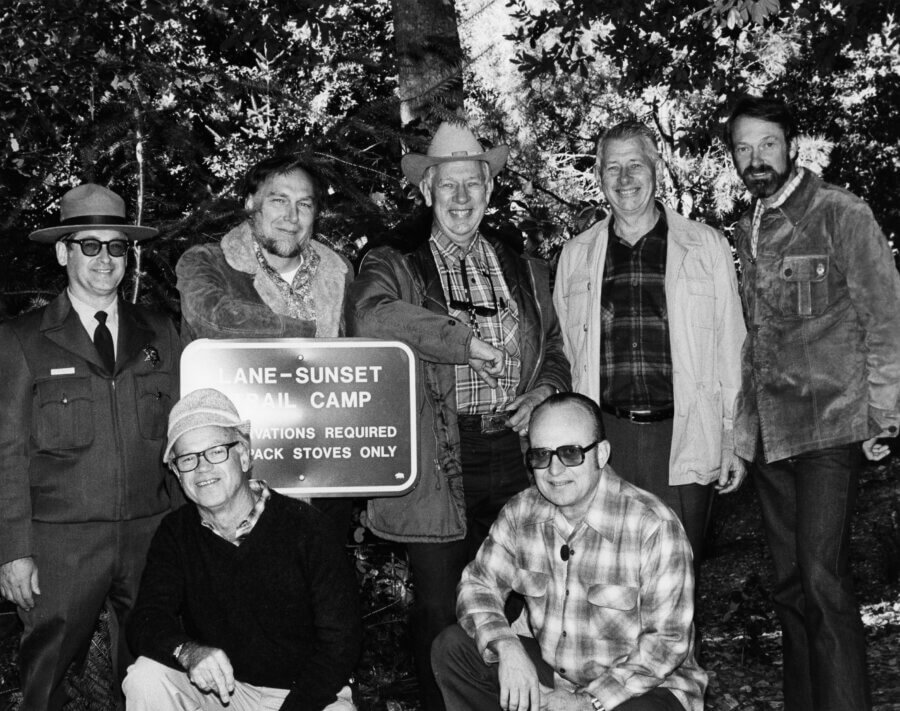 Seven people, some standing and some kneeling, pose together outdoors beside a sign reading Lane-Sunset Trail Camp. They are dressed in outdoor and winter clothing, smiling at the camera. Trees and foliage are in the background.