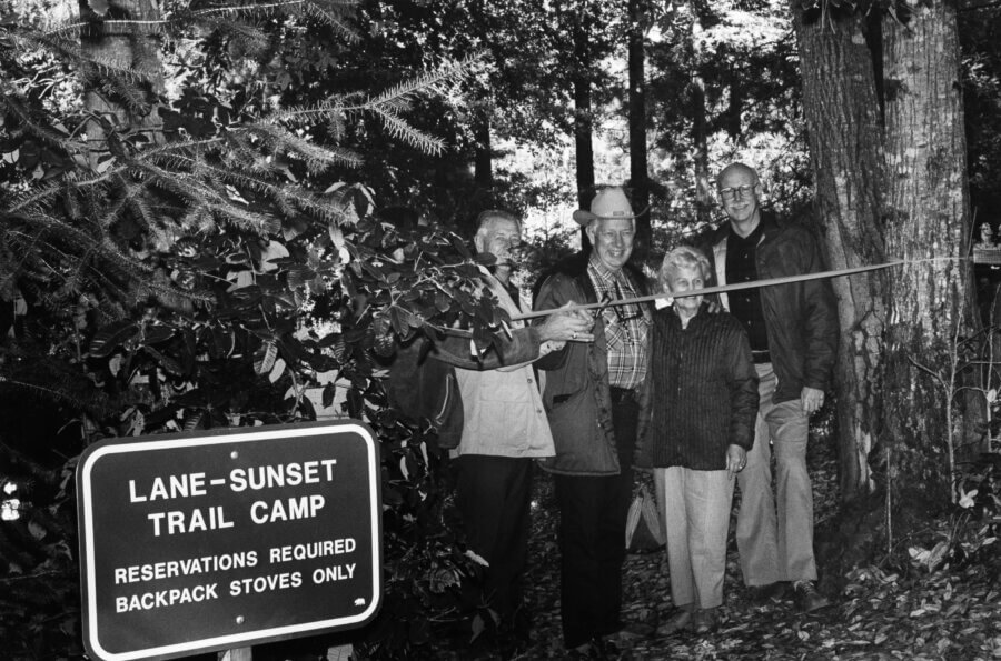 Four people stand smiling in a wooded area as one cuts a ribbon stretched across a trail. Nearby, a sign reads LANE-SUNSET TRAIL CAMP RESERVATIONS REQUIRED BACKPACK STOVES ONLY.