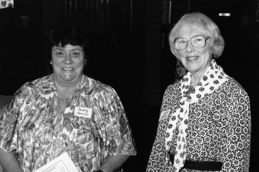 Two people smiling at the camera. Both are wearing glasses; one has a floral shirt and a name tag, the other wears a patterned dress with a scarf. The background is dark and indistinct.