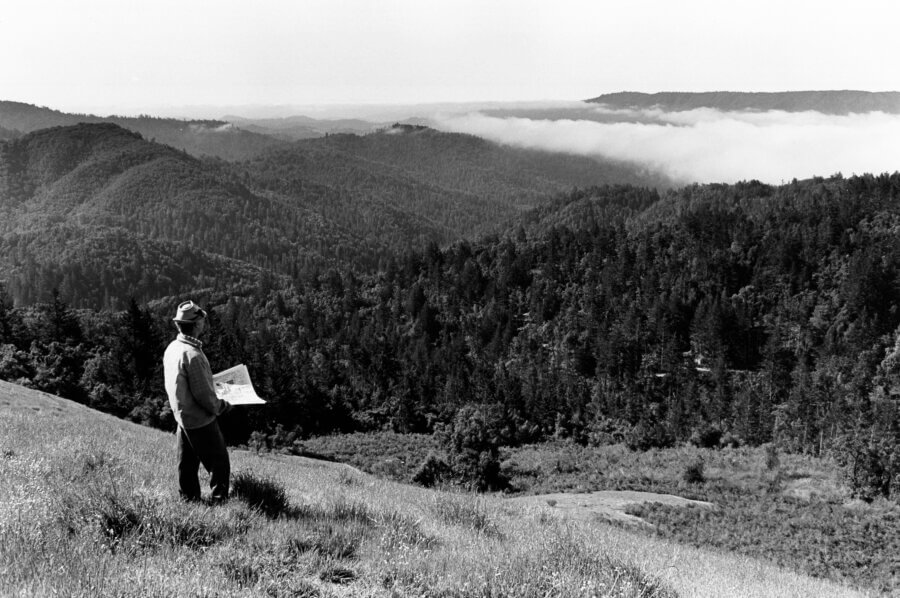 A person stands on a grassy hilltop, holding a map and looking out over a vast landscape of tree-covered hills and distant fog under a partly cloudy sky.