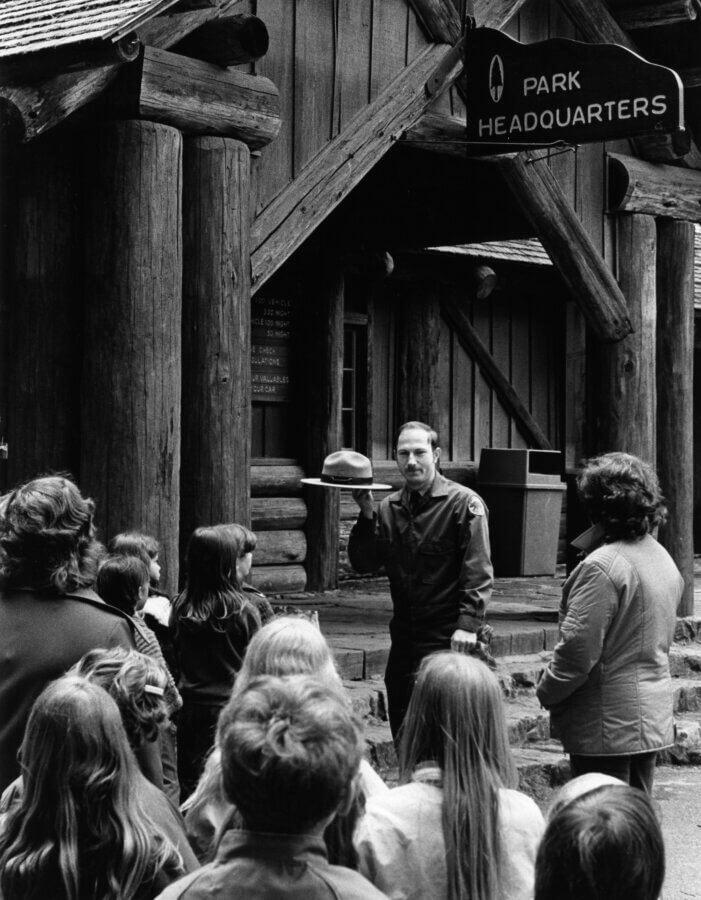 A park ranger holding a hat speaks to a group of children and adults outside a rustic wooden building with a Park Headquarters sign.