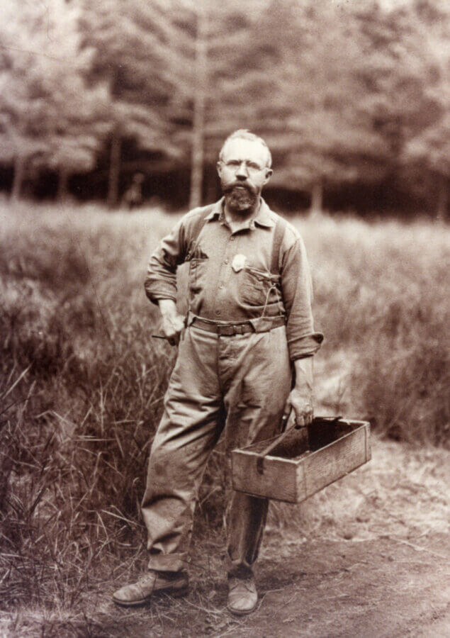 A bearded person stands outdoors in tall grass, wearing work clothes and holding a wooden box in one hand. Trees and blurred foliage are visible in the background.