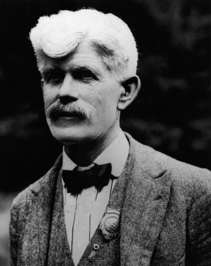A black-and-white portrait of an older person with white hair and a mustache, wearing a textured suit jacket, bow tie, and a round badge pinned to his vest. The background is out of focus.