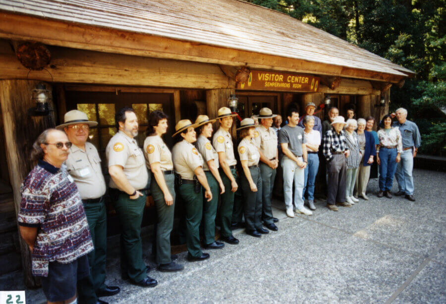 A group of people, including park rangers in uniform and civilians, stand in a line outside a wooden visitor center building surrounded by trees. The building has a sign reading Visitor Center above the entrance.