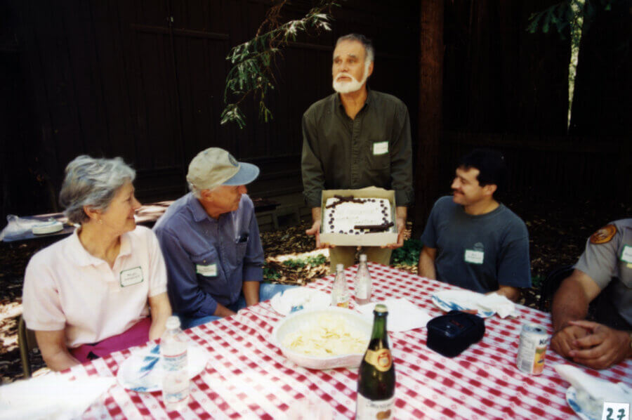 Four people sit at a picnic table with a red-checkered tablecloth, while another person stands holding a decorated cake. Food, drinks, and name tags are visible. The scene is outdoors, with trees and a wooden building in the background.