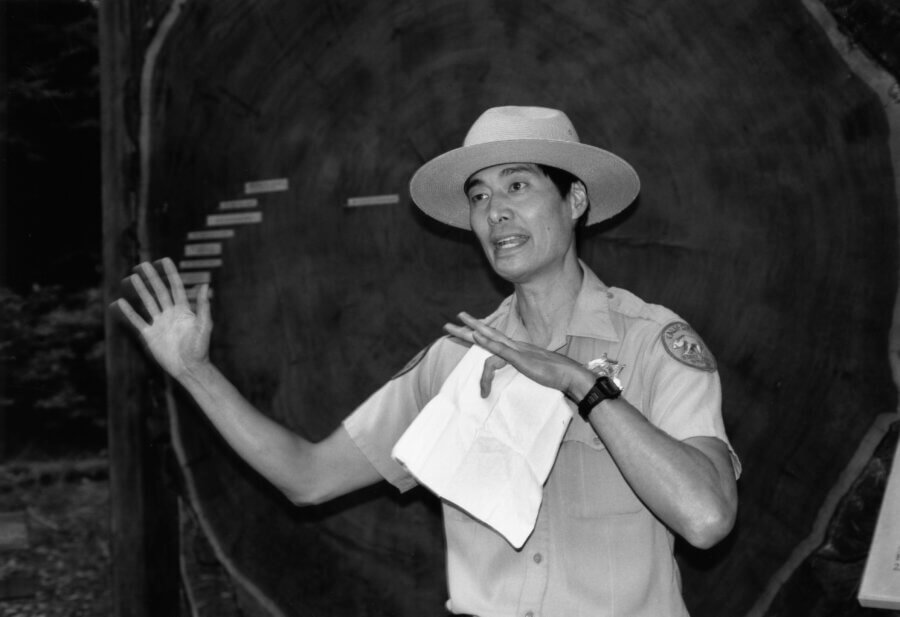A park ranger in uniform and hat gestures while speaking in front of a large cross-section of a tree trunk, with historical date markers visible on the wood.