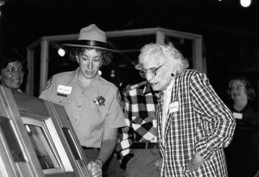 A park ranger shows an exhibit to a person in a plaid jacket, while several other people observe and smile in the background. The scene appears to take place indoors.