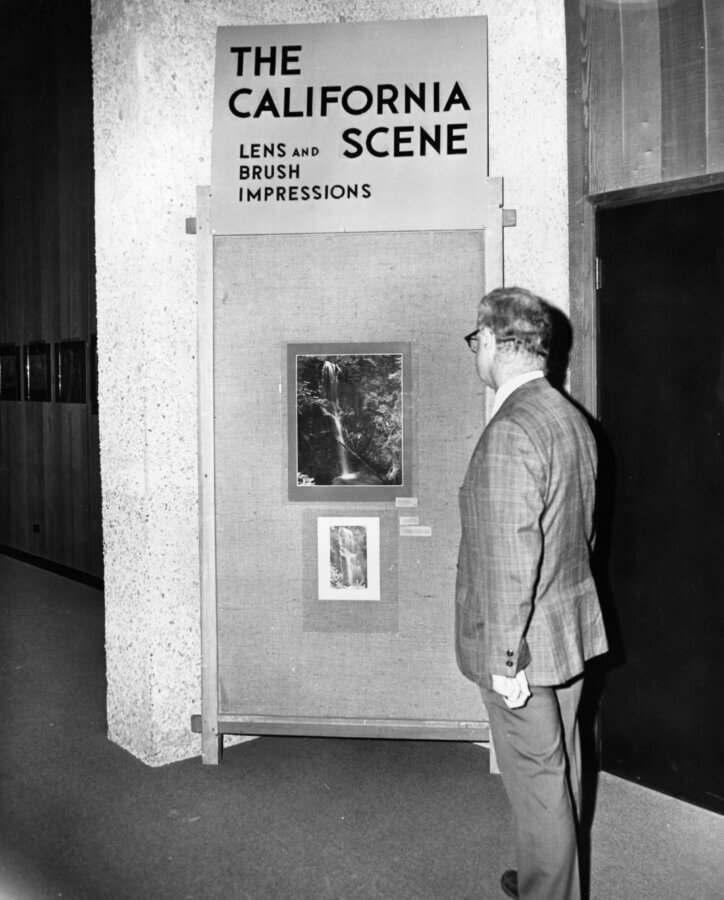 A person in a suit looks at a display titled The California Scene: Lens and Brush Impressions, featuring photographs of a waterfall, mounted on a fabric-covered board in a gallery setting.