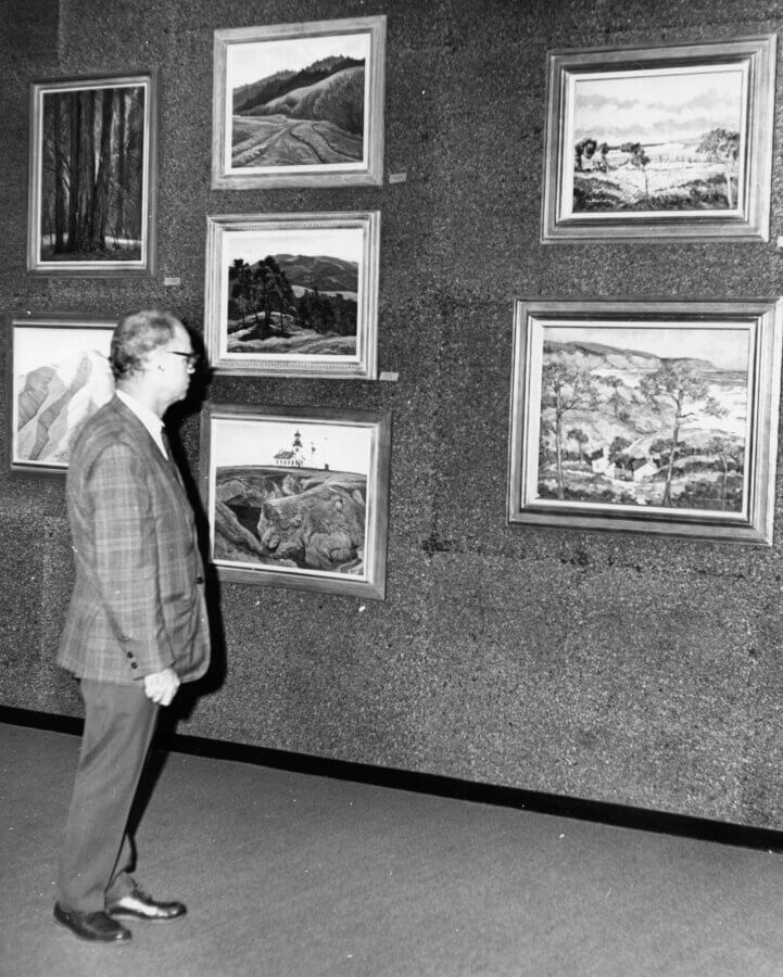 A person in a plaid jacket and glasses stands indoors, looking at a wall display of six framed landscape paintings. The artworks depict forests, mountains, and scenic outdoor views.