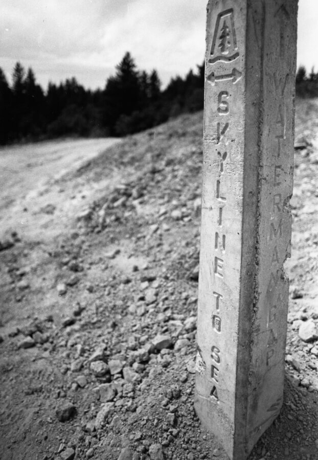 A weathered concrete trail marker stands on rocky ground, engraved with the words SKYLINE TO SEA and an emblem of a tree. Trees and a cloudy sky are visible in the background.