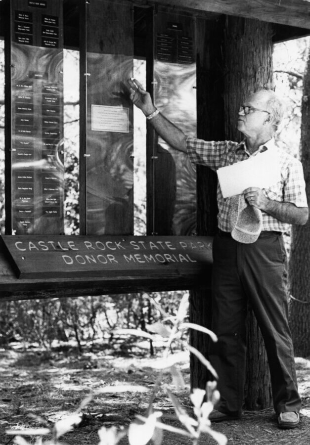 A person stands at a wooden display titled Castle Rock State Park Donor Memorial, pointing at a plaque while holding papers in one hand. Trees and forest surroundings are visible in the background.