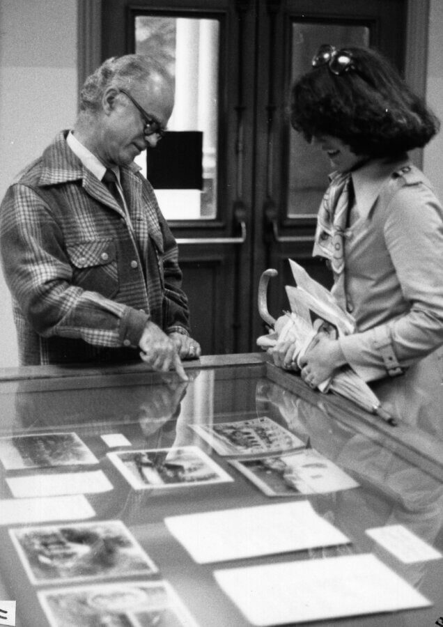 A person points at photographs in a glass display case while another person holding papers listens attentively. They are indoors, standing in front of double wooden doors.
