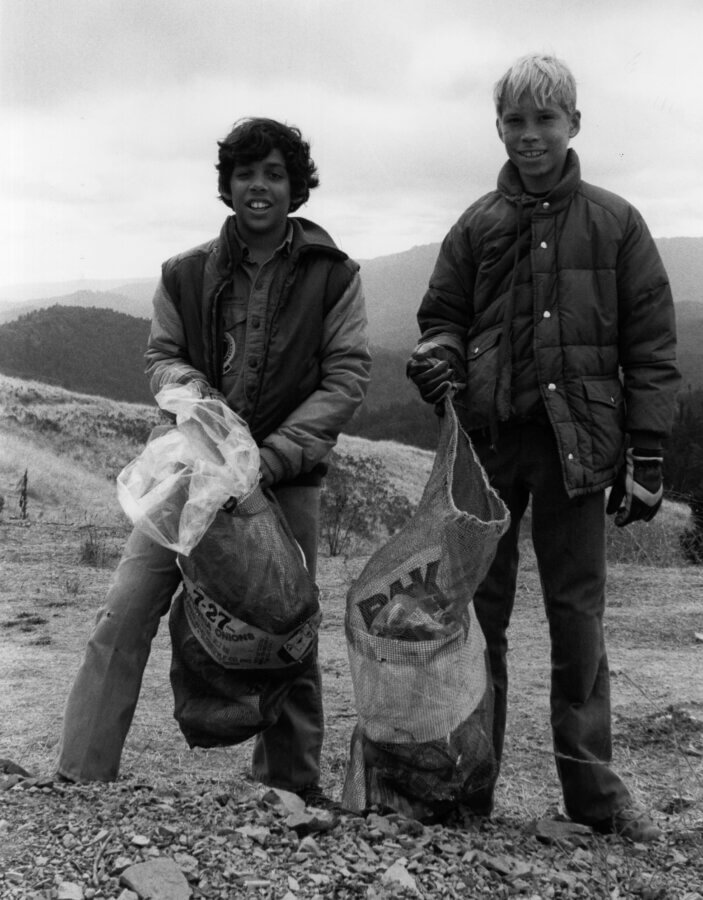 Two children stand outdoors on a rocky hillside, each holding a large bag full of collected items. Both wear jackets and gloves, with mountains and cloudy skies visible in the background.