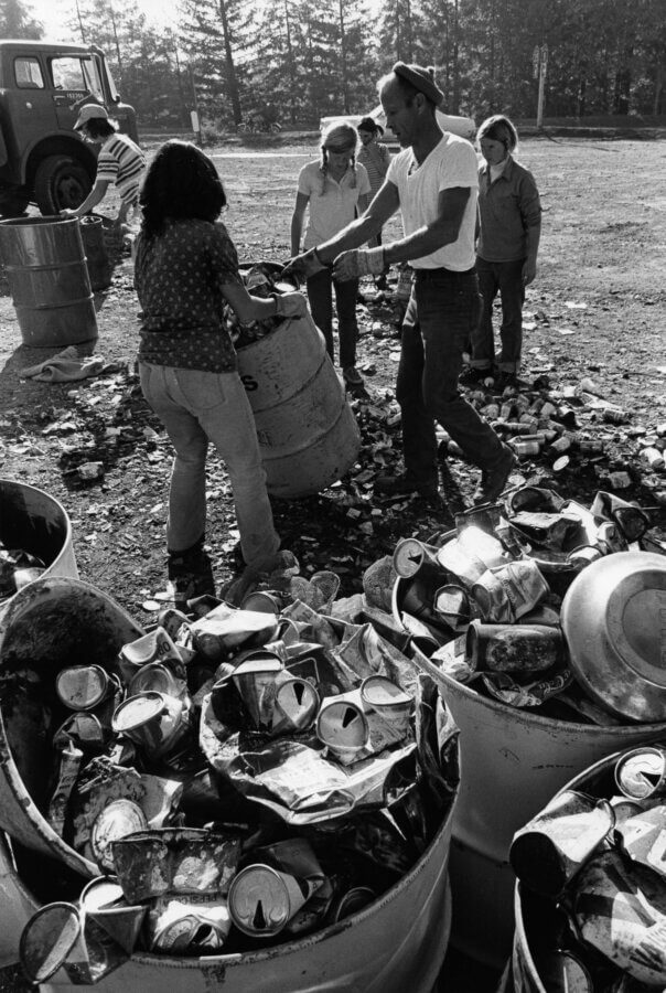 A group of people outdoors sorts and collects empty, crushed metal cans into large barrels, with more barrels full of cans in the foreground and a truck visible in the background.