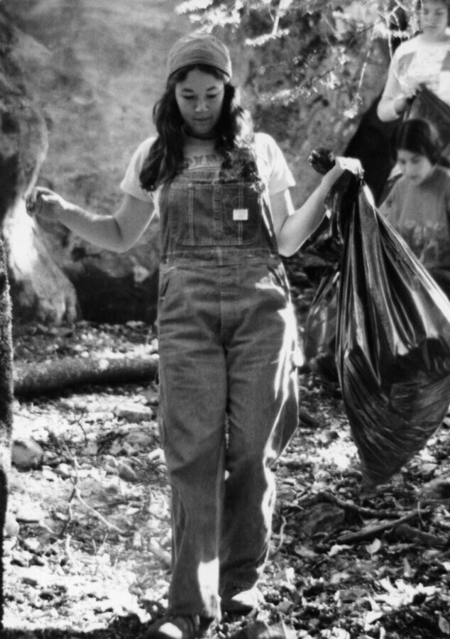 A person in overalls and a cap walks outdoors, holding a large trash bag and surrounded by trees and fallen leaves, participating in a cleanup effort.