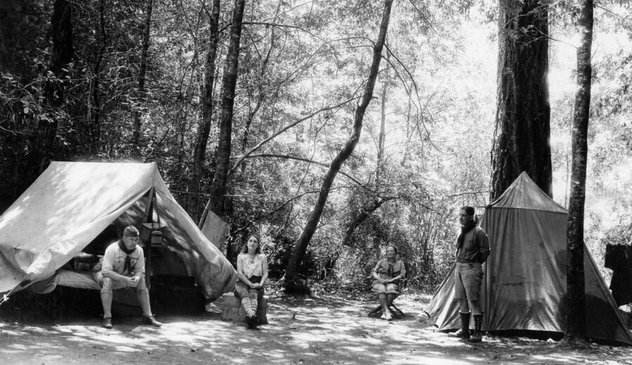 Four people sit and stand around tents in a wooded campsite. Sunlight filters through tall trees. One person sits at the entrance of a tent, another sits nearby, and two others are standing and sitting on the right.