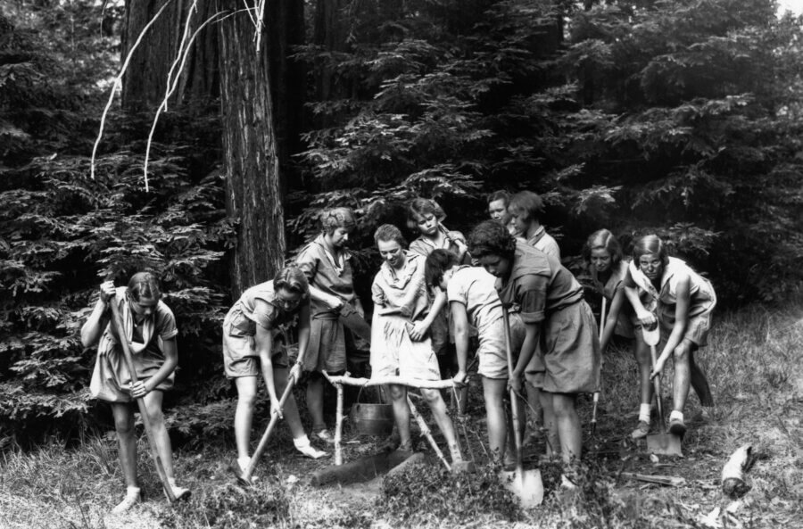 A group of people in shorts and shirts stand in a circle in a wooded area, digging in the ground with shovels, surrounded by tall trees and dense foliage.