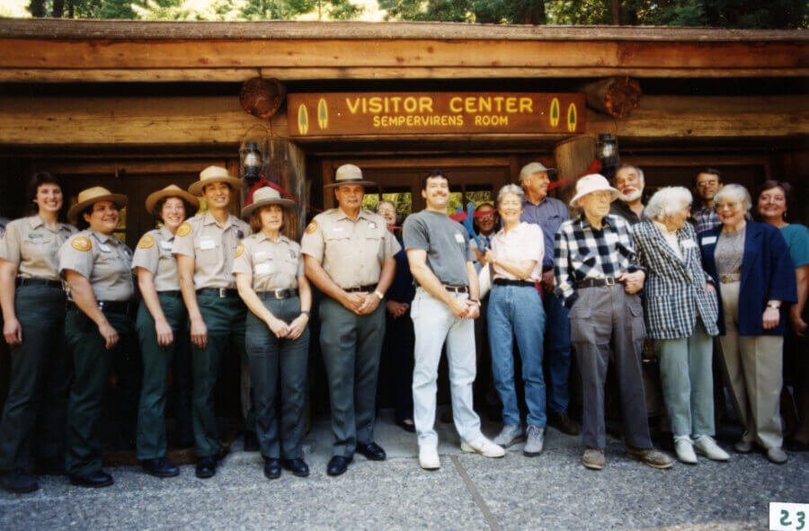 A group of park rangers and visitors pose smiling in front of a wooden Visitor Center with a sign reading Visitor Center Sempervirens Room on a sunny day.