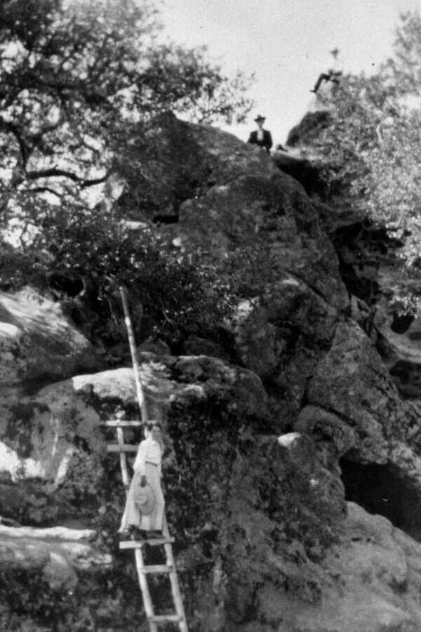 A person stands on a wooden ladder leaning against a rocky cliff, while two people sit at the top of the large rocks surrounded by trees.