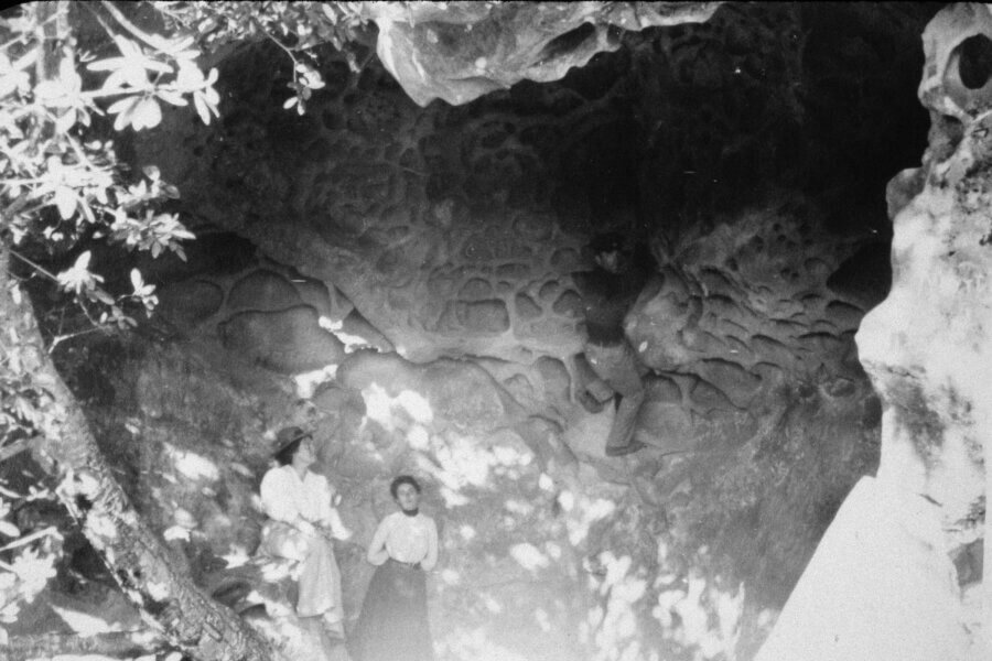 Two people stand beneath a large, textured rock overhang while another person climbs the rock face above them. Sunlight filters through tree branches, casting dappled shadows on the scene.