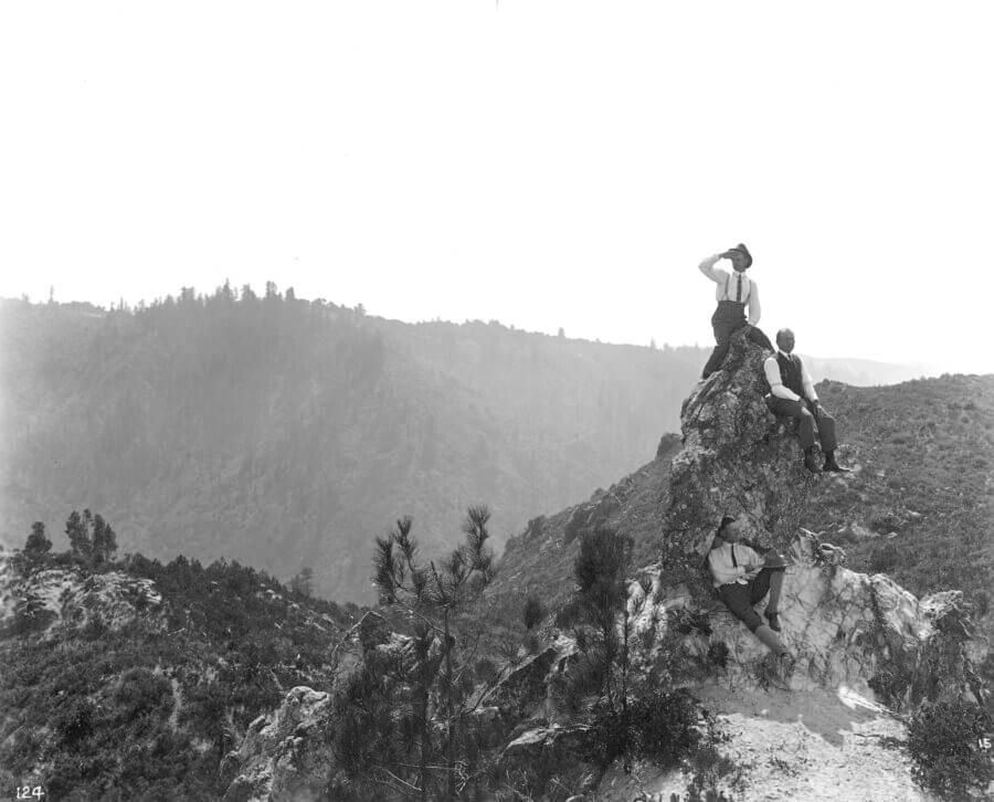 Three people in early 20th-century attire sit and stand on a rocky hillside, looking out over a vast, mountainous landscape with sparse trees under a clear sky.