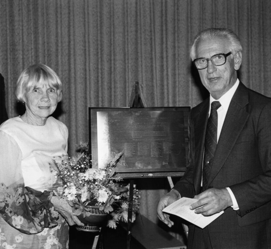A person holding a bouquet of flowers stands next to another person in a suit and tie. Behind them is a plaque displayed on an easel, and a curtain is visible in the background.