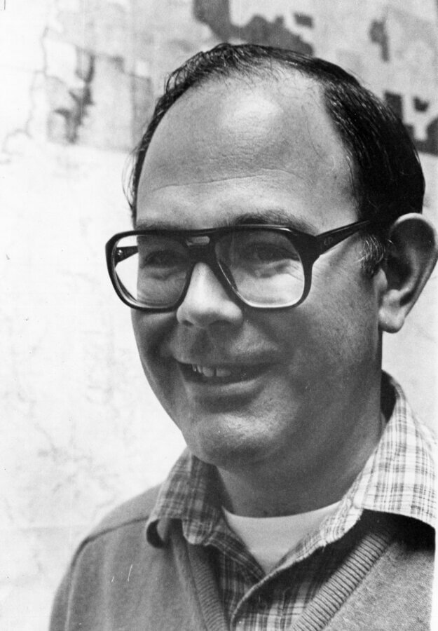 A person with glasses smiles while wearing a plaid shirt and sweater.